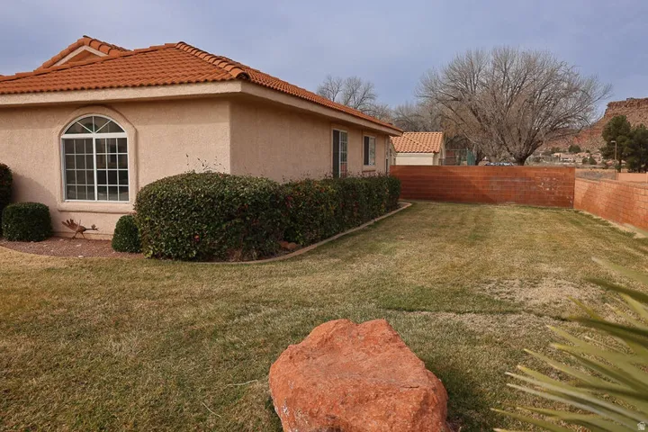 Expansive grassy yard featuring a decorative rock, a privacy wall, established shrubbery, and a stucco exterior with a tiled roof
