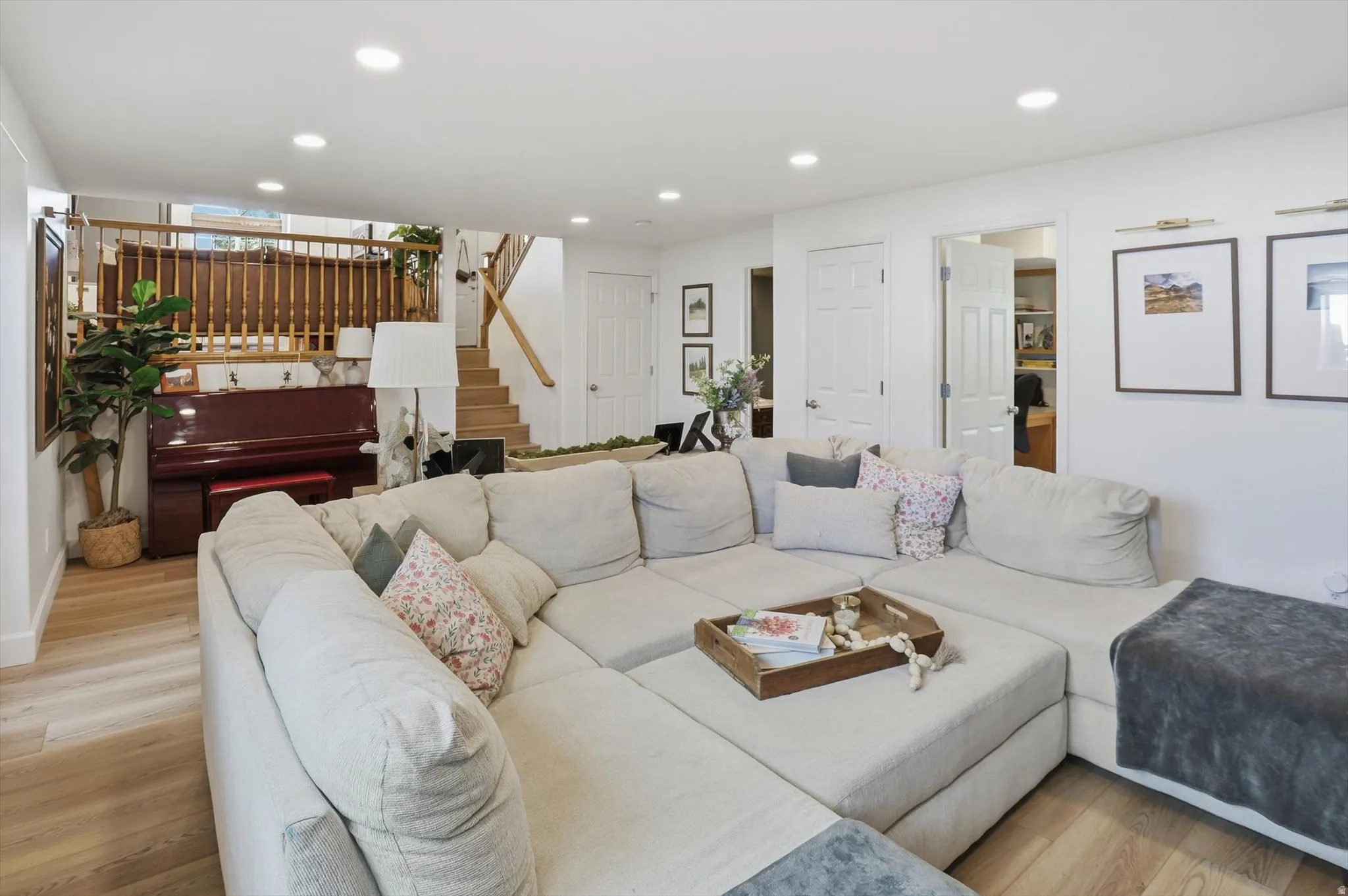 Spacious living area featuring light wood-finish flooring, recessed lighting, white walls, and a visible staircase with wood railing