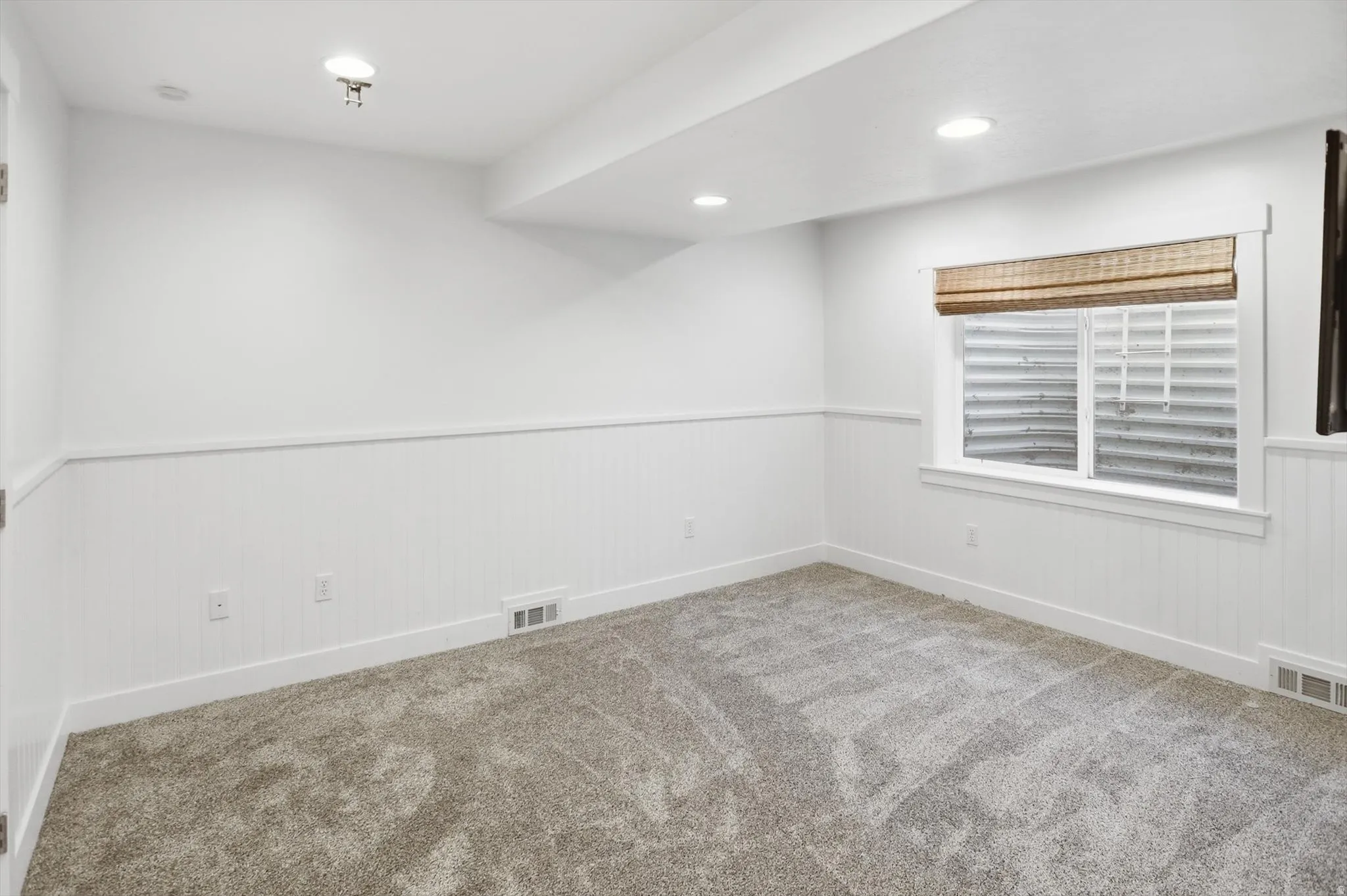 Recessed lighting illuminates this space featuring wainscoting, light-toned carpeting, and a window with a woven shade