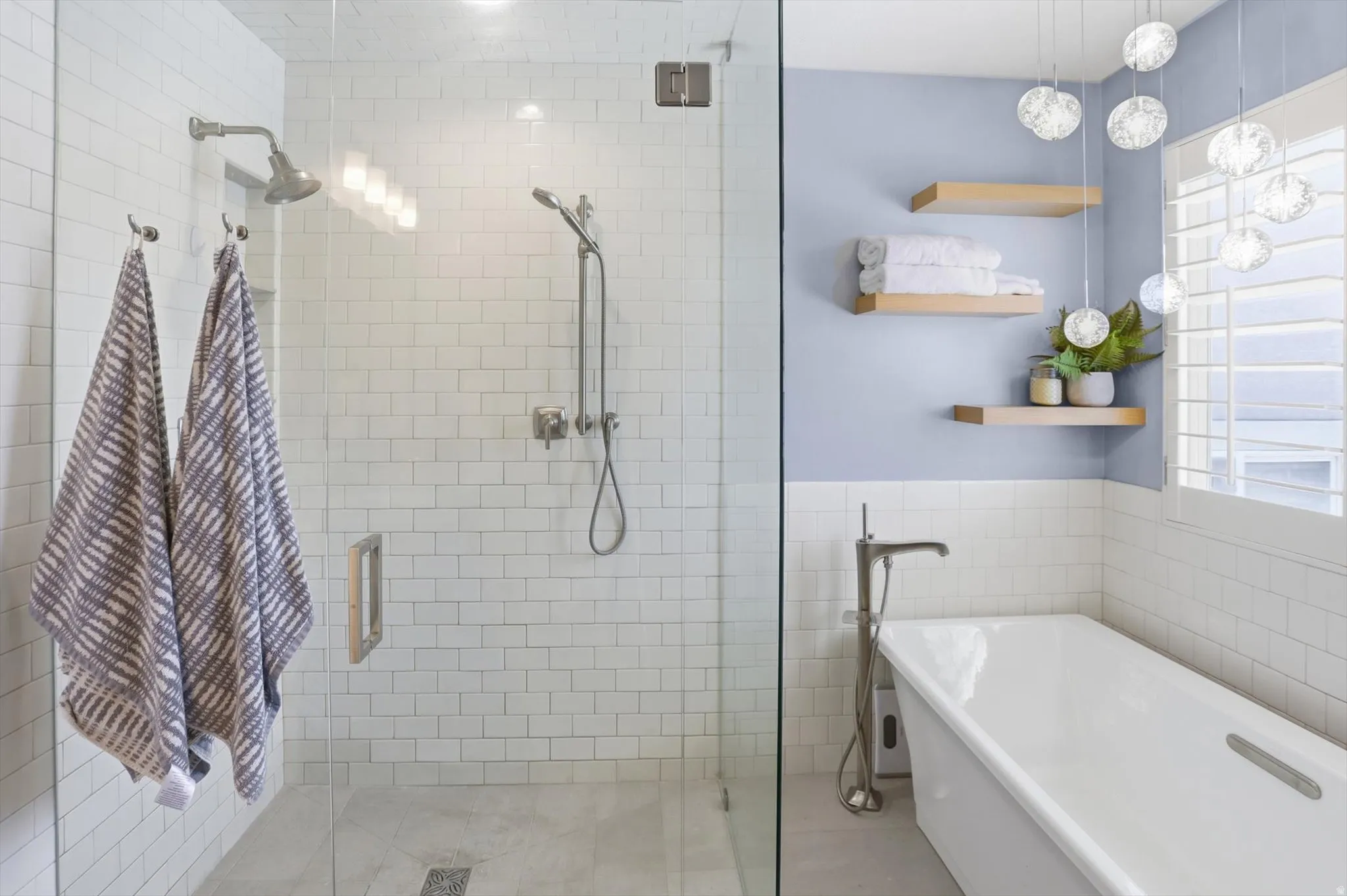 Spacious bathroom featuring a glass-enclosed shower with subway tile surround, a freestanding soaking tub with a floor-mounted faucet, and floating wood shelves