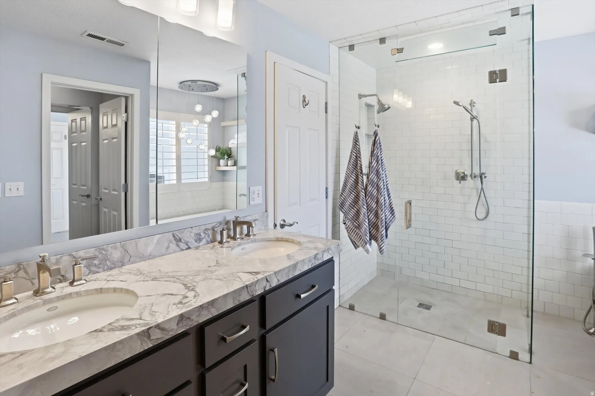 Spacious bathroom featuring a frameless glass shower enclosure with white subway tile, a dual vanity with a patterned stone countertop, and light blue wall paint