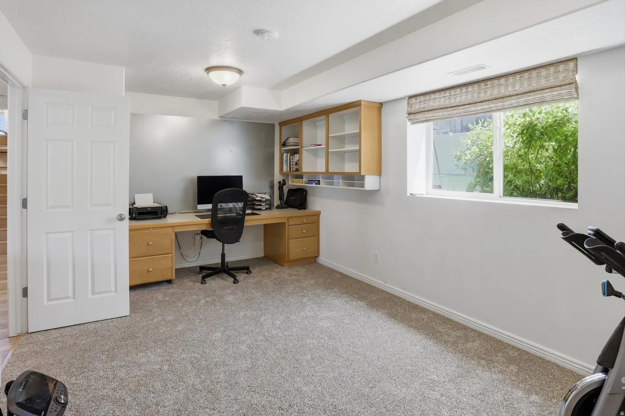 Carpeted room featuring a built-in wood-finish desk with integrated cabinetry and shelving, a window with a Roman shade, and an overhead flush-mount light fixture