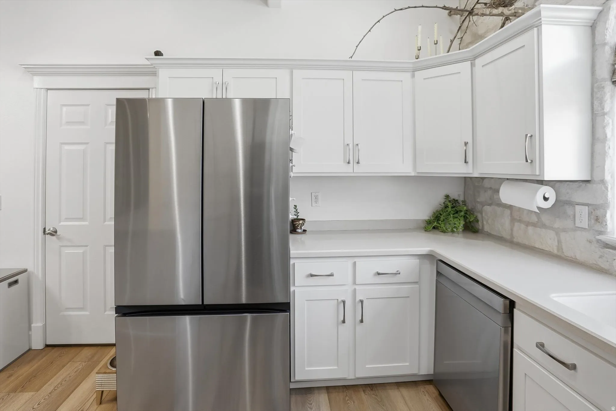 Stainless steel French door refrigerator, white shaker-style cabinetry with chrome hardware, light-toned countertops, stone backsplash, and wood-finish flooring