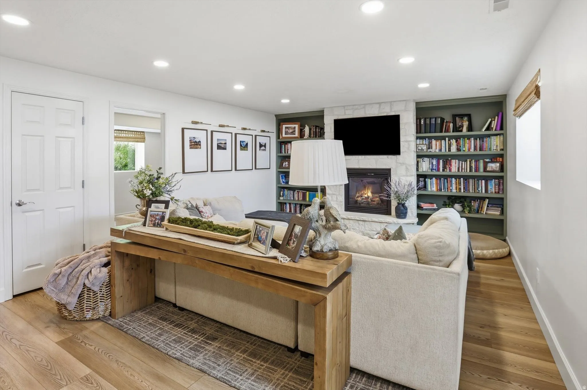 Living area featuring a stone-surround fireplace with built-in shelving, wood-finish flooring, recessed lighting, and a white six-panel door