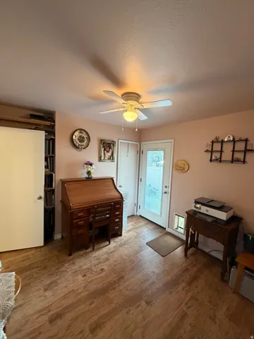 Wood-finish flooring throughout, featuring a ceiling fan with integrated lighting, a white exterior door with glass insert, and built-in shelving