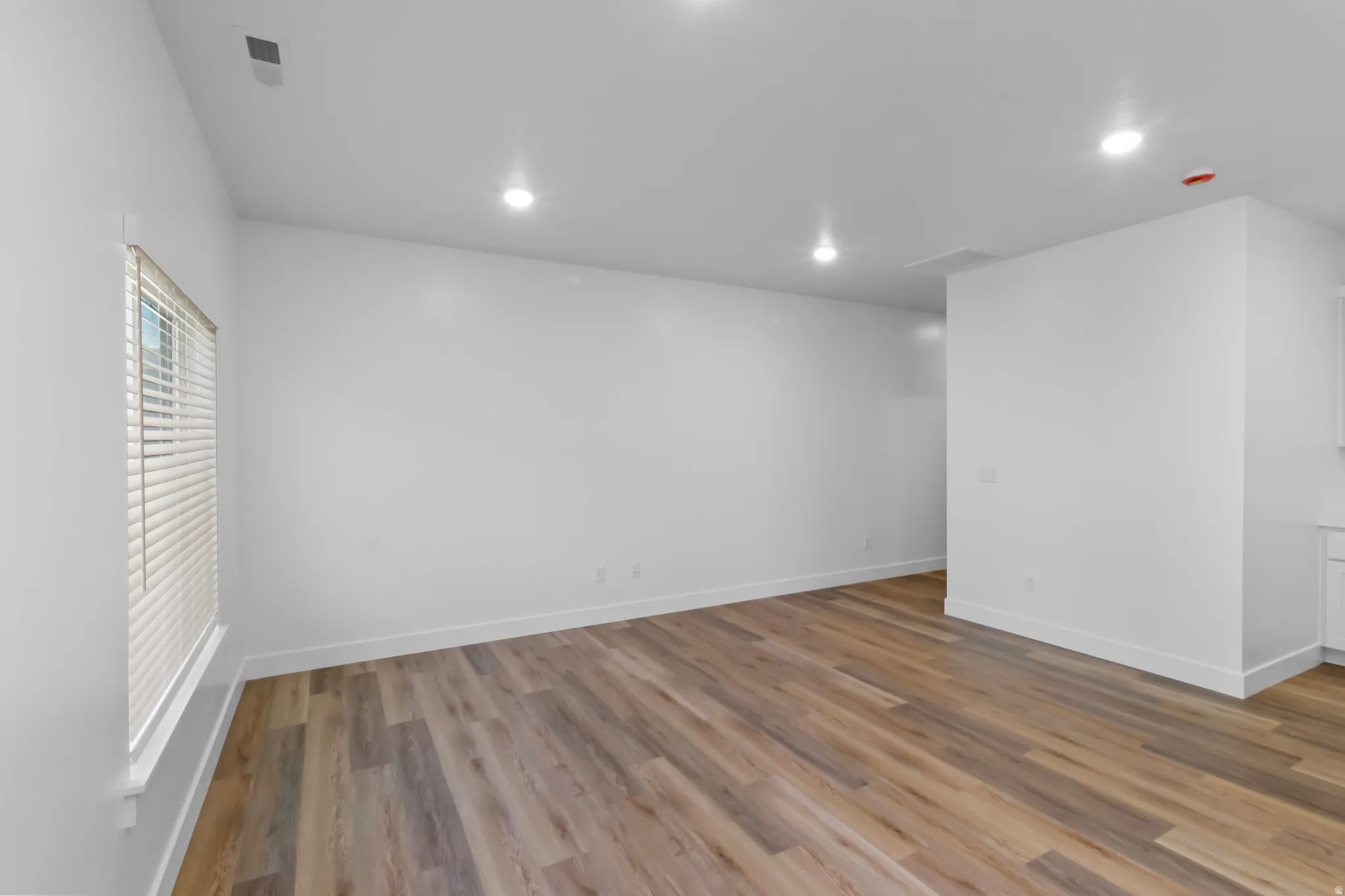 Spacious interior with neutral white walls, wood-finish flooring, recessed lighting, and a single window with horizontal blinds