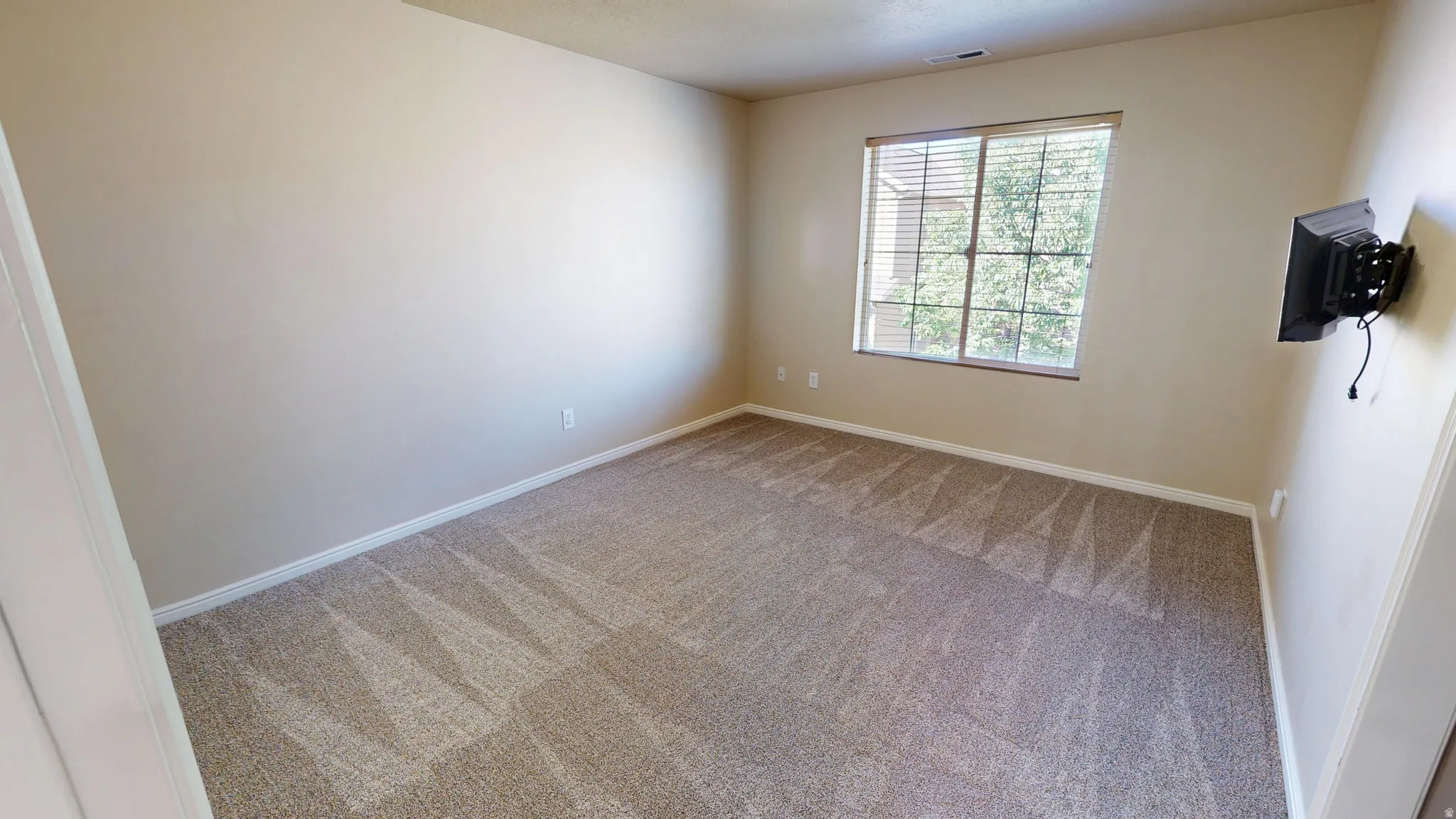 Spacious room featuring neutral wall tones, patterned carpet, a large window with horizontal blinds, and a mounted bracket
