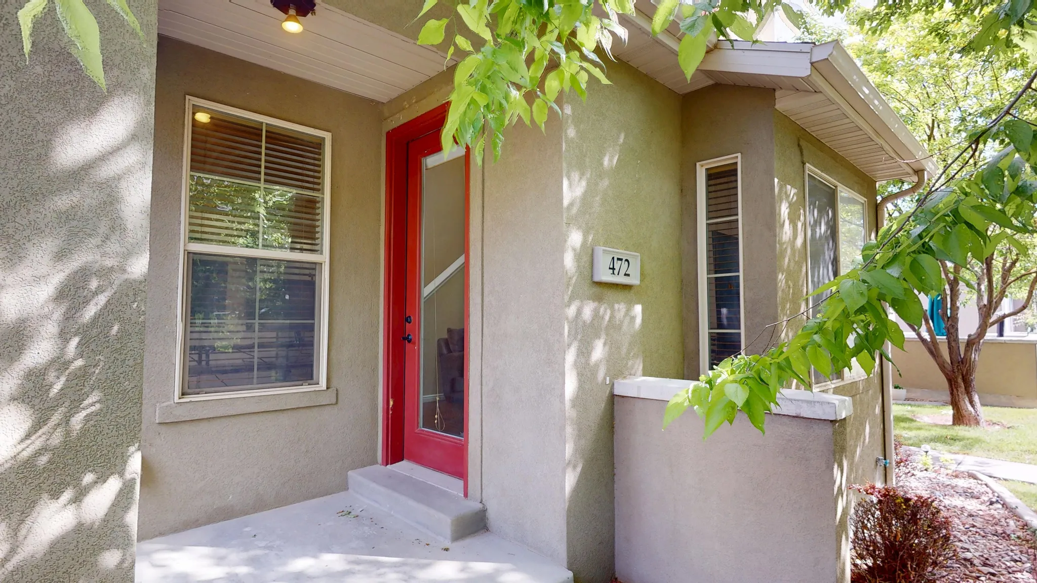 Stucco exterior featuring a prominent red entry door with sidelight, large window with blinds, and an illuminated overhead light fixture