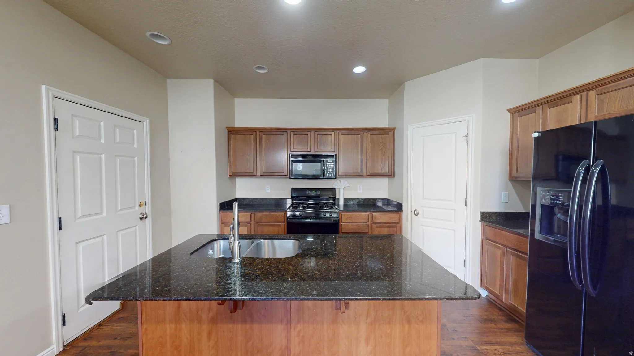 Kitchen featuring a central island with an undermount sink, dark speckled countertops, wood-finish cabinetry, a black range with an overhead microwave, and a black French door refrigerator