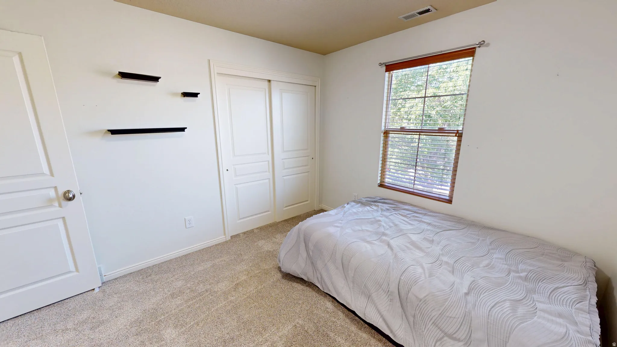 Bright interior space featuring a window with wood-finish blinds, light-toned carpet flooring, a white sliding closet door, a white interior door, and three wall-mounted black floating shelves