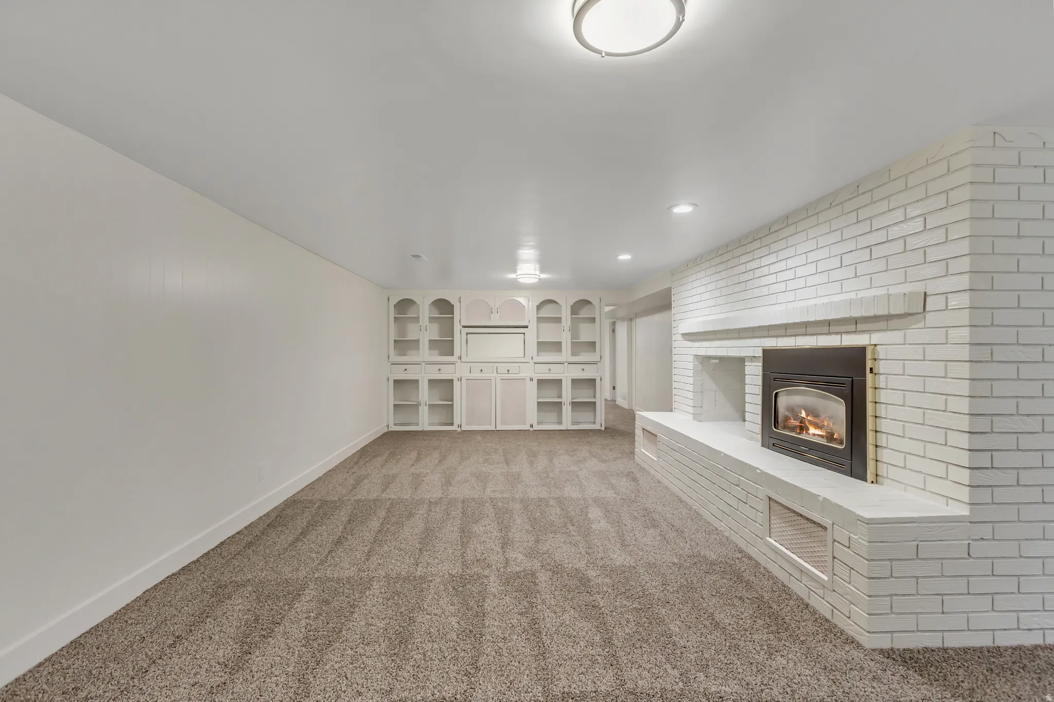 Spacious room featuring a white painted brick fireplace with a raised hearth, built-in shelving, recessed lighting, and neutral carpet