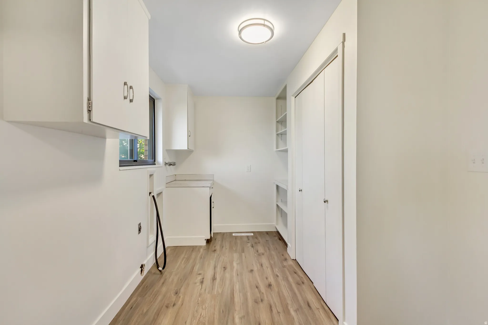 Utility room featuring wood-finish flooring, white cabinetry, a built-in countertop, a window, and a bifold door closet with integrated shelving