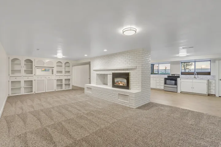 Living area with a white brick fireplace, built-in cabinetry, and neutral carpet, opening to a kitchen featuring light wood-finish flooring, white cabinetry, and stainless steel appliances
