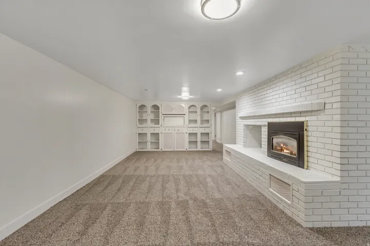 Spacious room featuring a white painted brick fireplace with a raised hearth, built-in shelving, recessed lighting, and neutral carpet