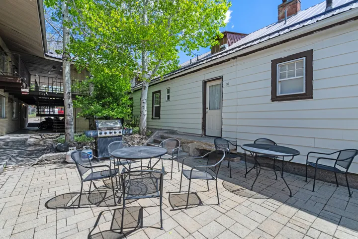 Paved patio area featuring light-toned paver stones, multiple mature trees, and a white-sided building with a brown-trimmed window and door