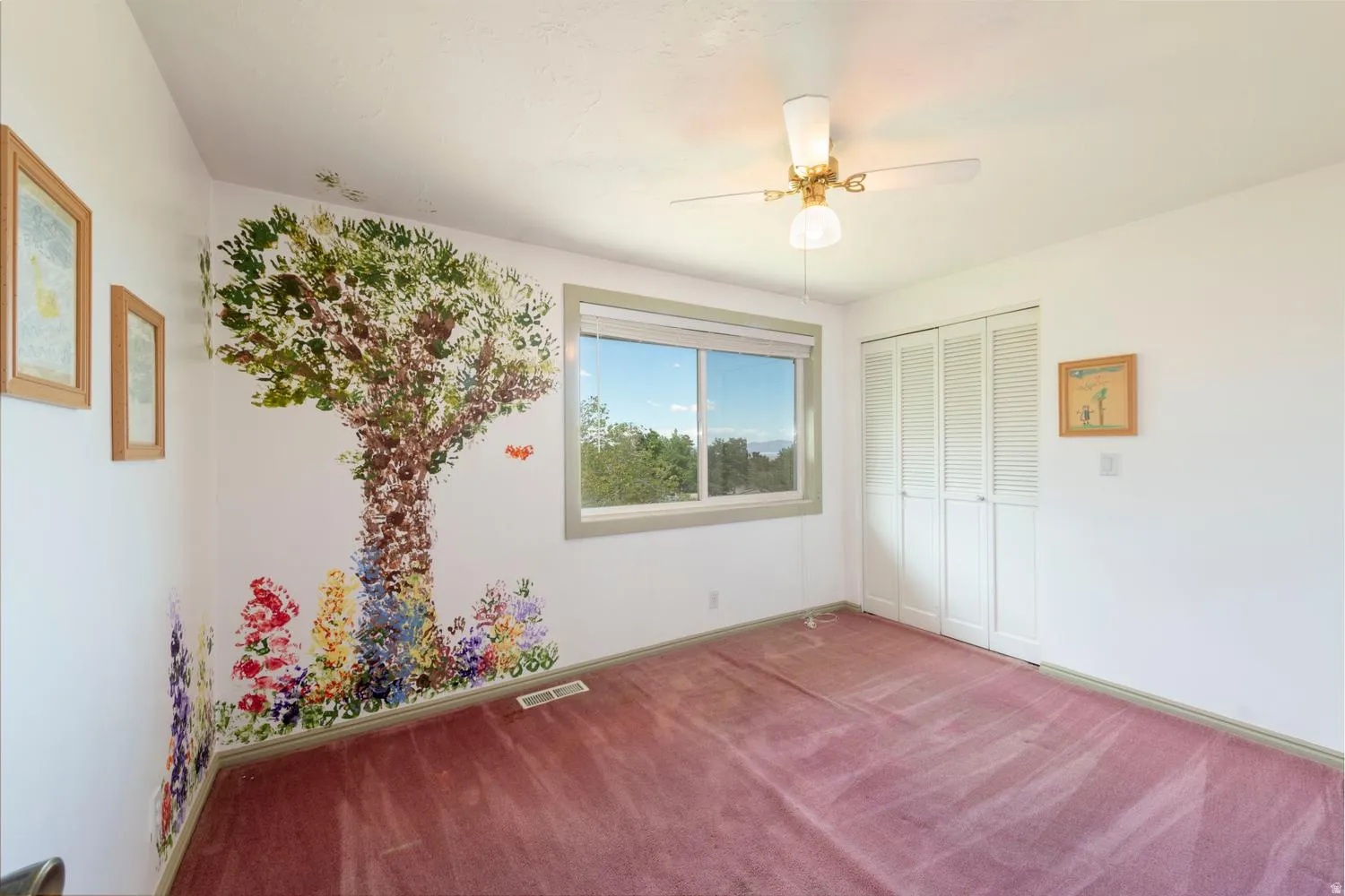 Room featuring a painted mural, a ceiling fan with light fixture, a window with exterior scenery, bi-fold closet doors, and carpet flooring