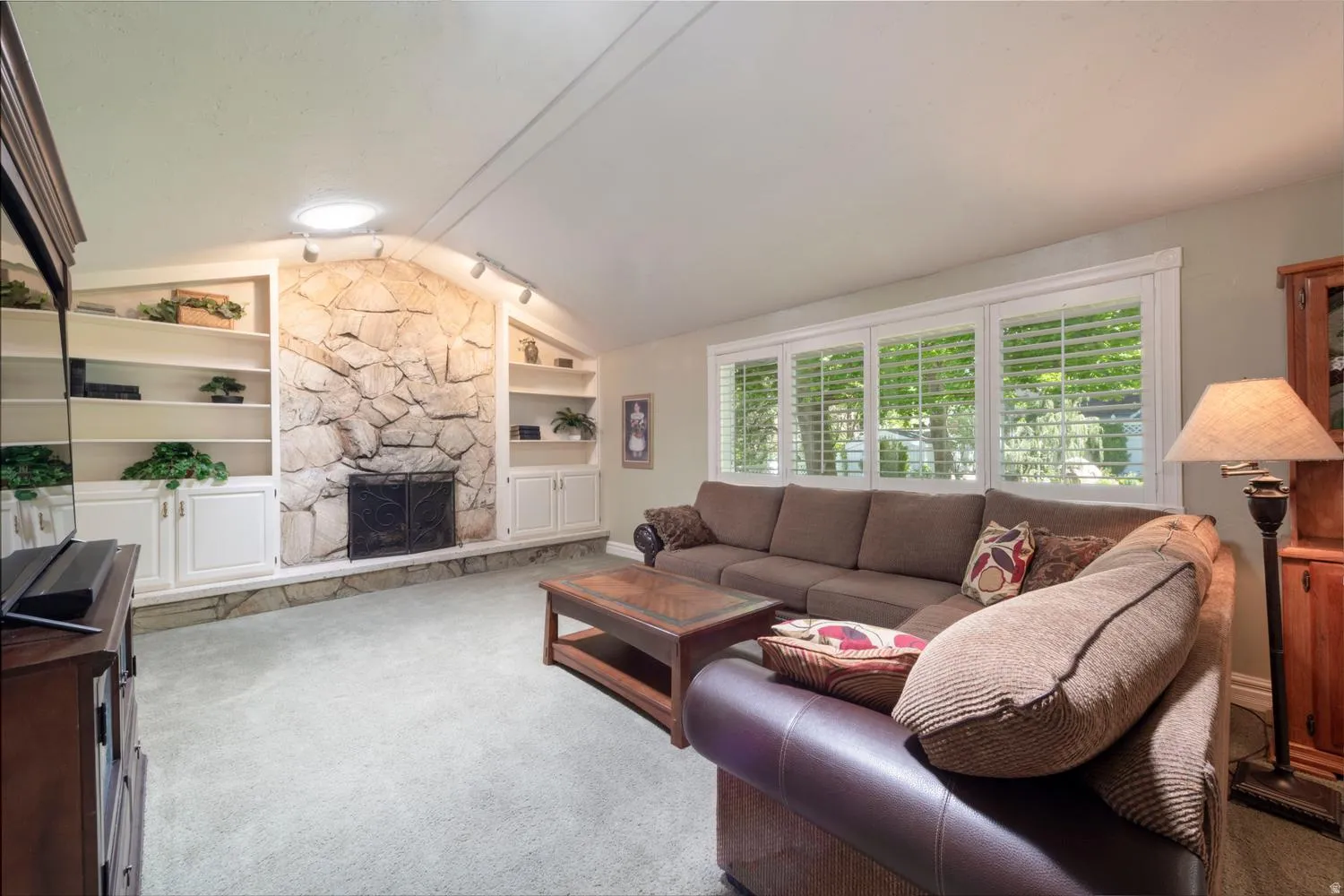 Living area featuring a stone fireplace with a black metal screen, flanked by built-in shelving and cabinetry, and multiple windows with plantation shutters