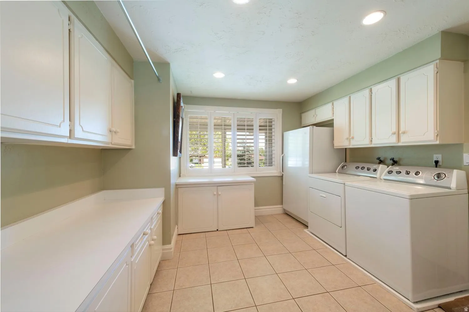 Spacious utility room featuring extensive white cabinetry, ample countertop space, a window with white plantation shutters, tiled flooring, and recessed lighting