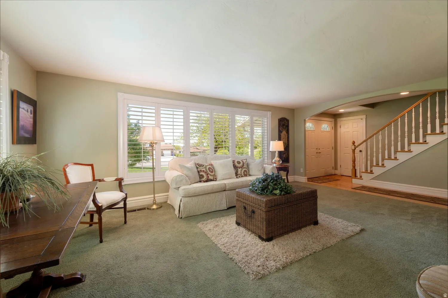 Light-filled living area featuring a large window with plantation shutters, plush green carpeting, and a grand staircase with white balusters and a wood handrail