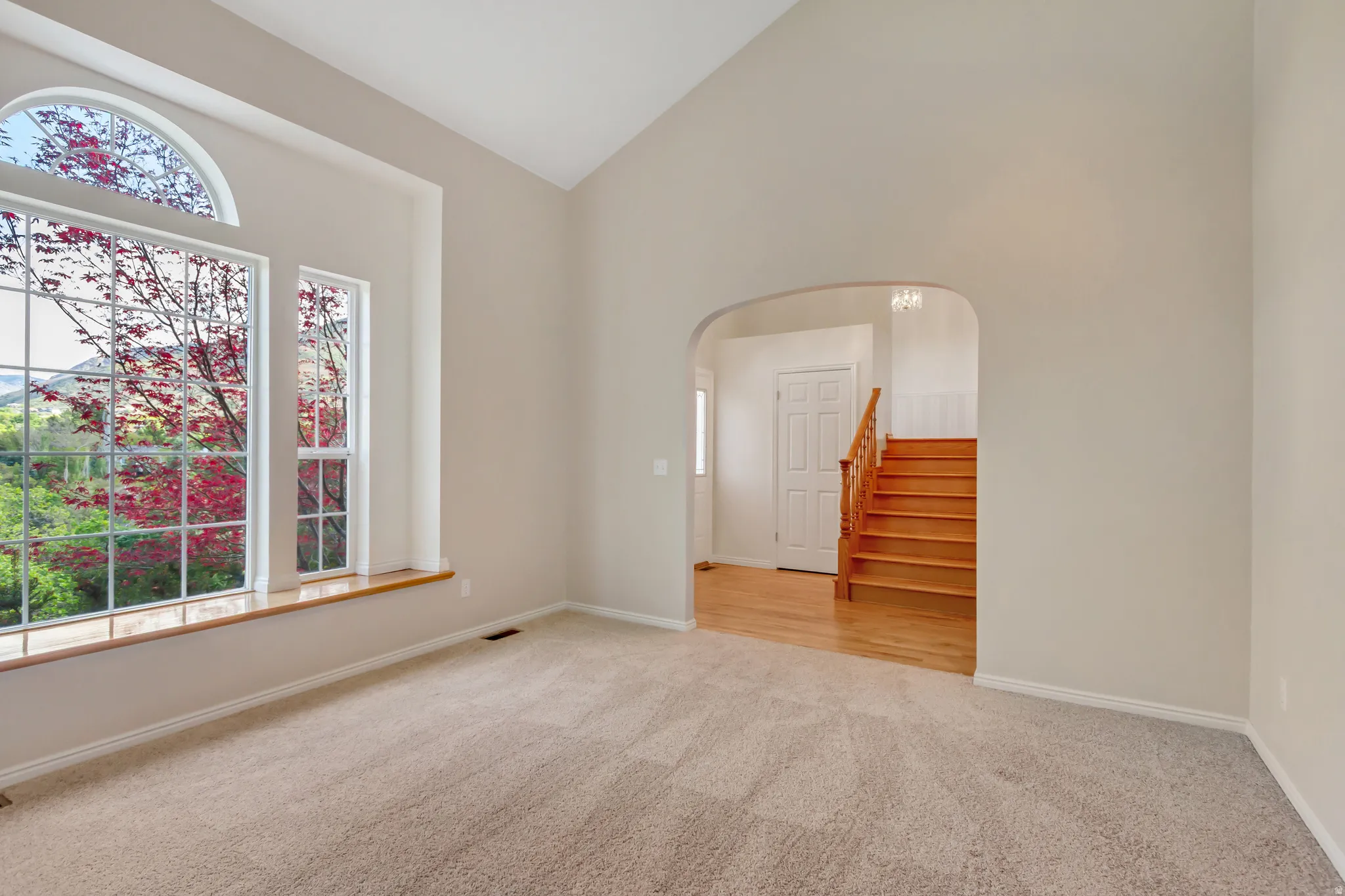 Vaulted ceiling room featuring large arched window, additional vertical window, light-toned carpet flooring, and a wide wooden window sill