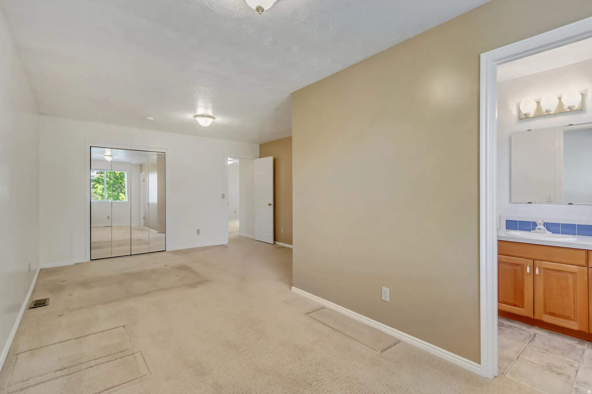 Neutral-toned room featuring a mirrored closet, ceiling-mounted light fixtures, and a doorway revealing a partial bathroom vanity with a mirror and light bar