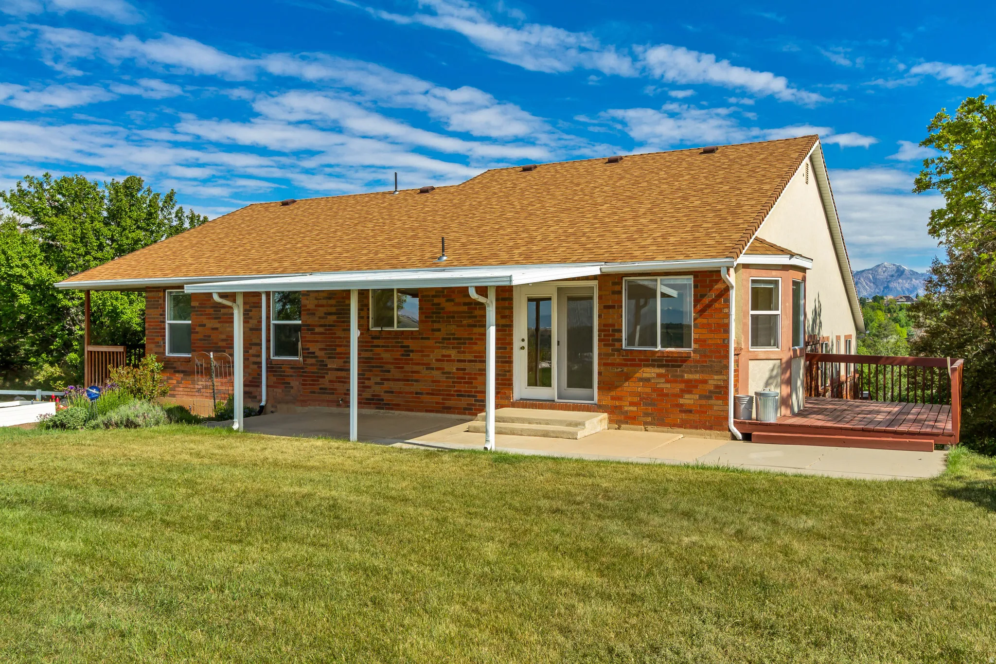 Expansive backyard featuring a manicured lawn, red brick exterior, brown shingle roof, covered patio, and an elevated wood deck