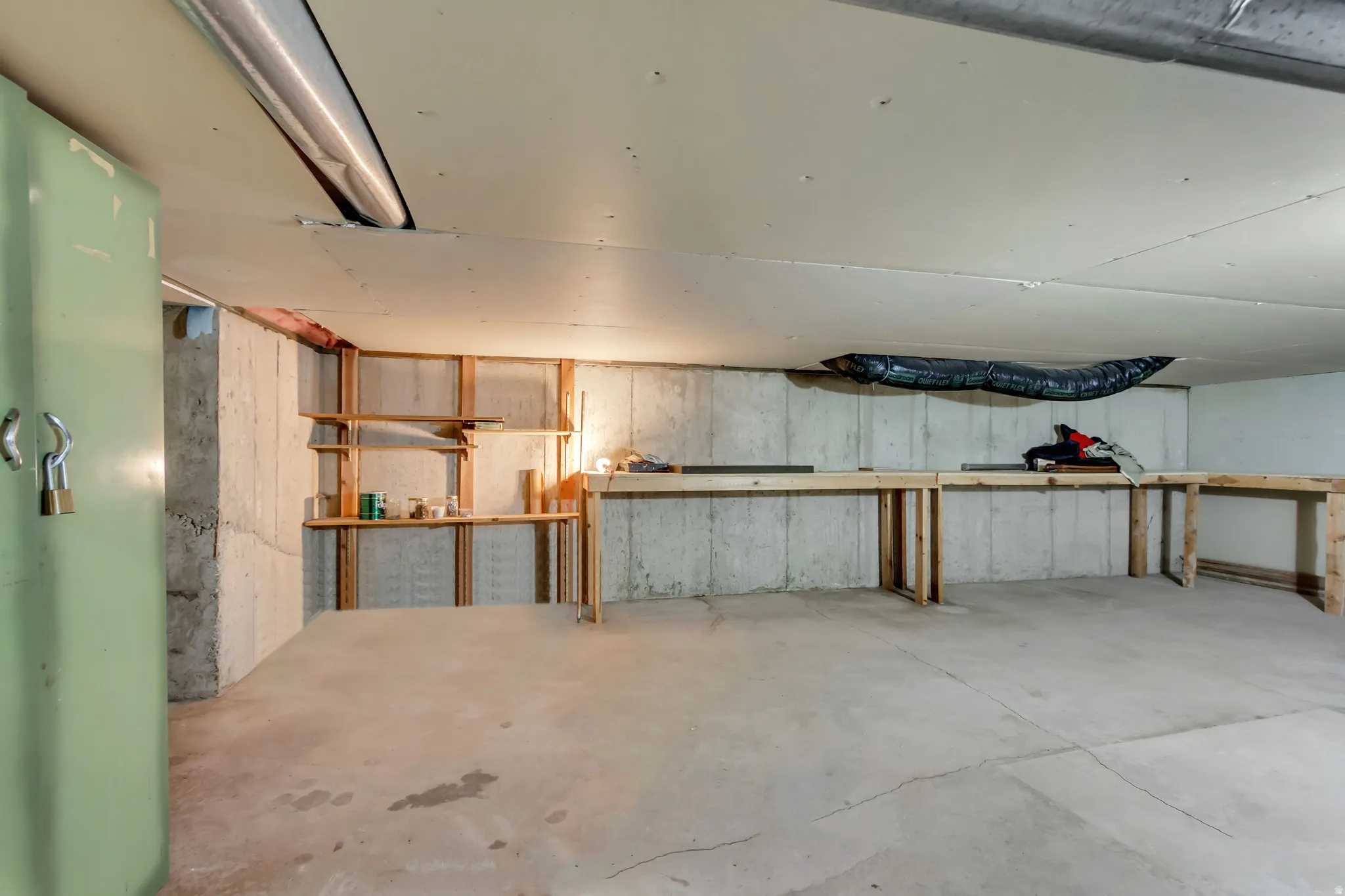 Unfinished basement featuring concrete walls and flooring, exposed ceiling joists, built-in shelving, and a long wooden workbench