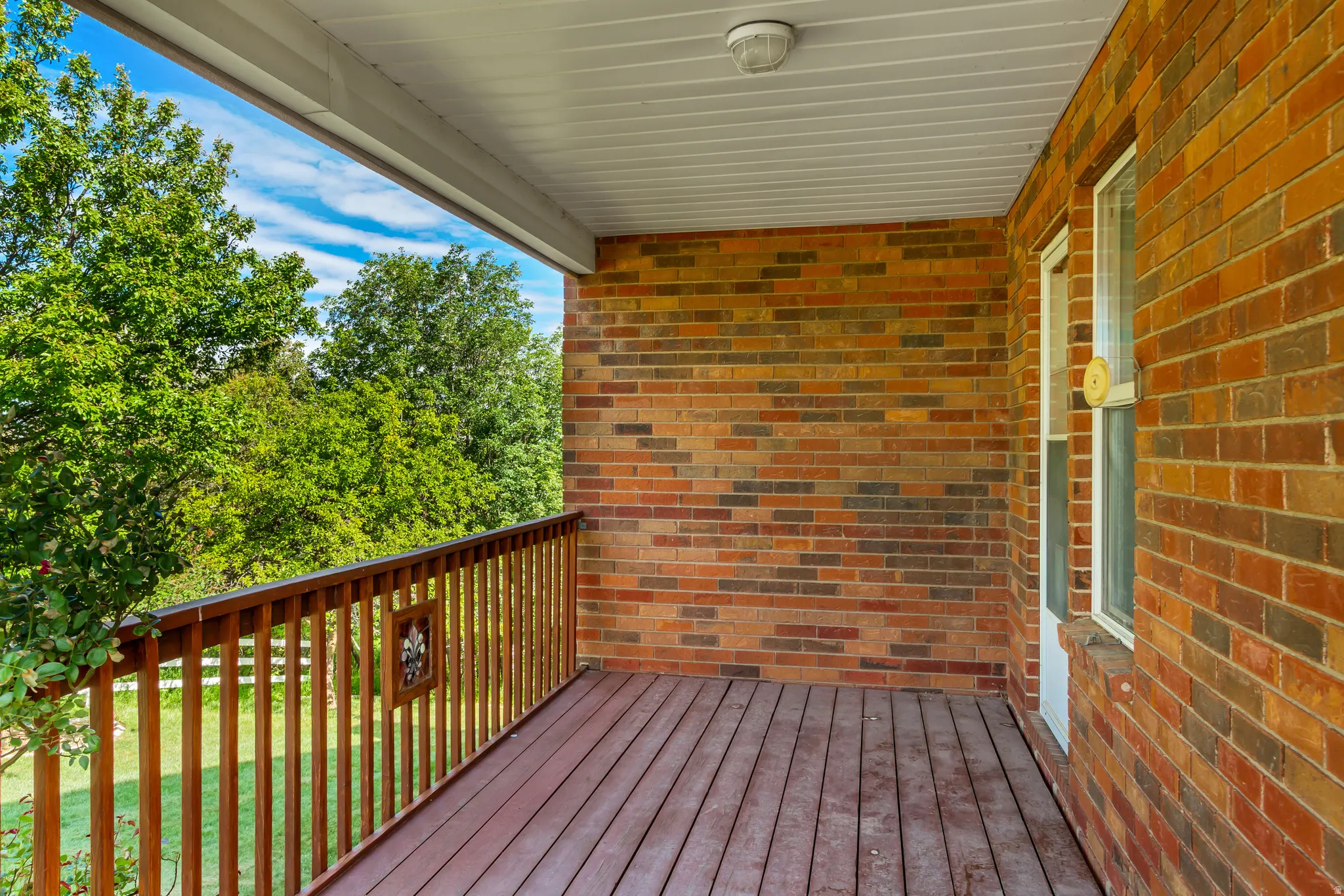 Covered outdoor deck with wood-finish flooring, featuring a wood railing with decorative accents and a brick exterior wall