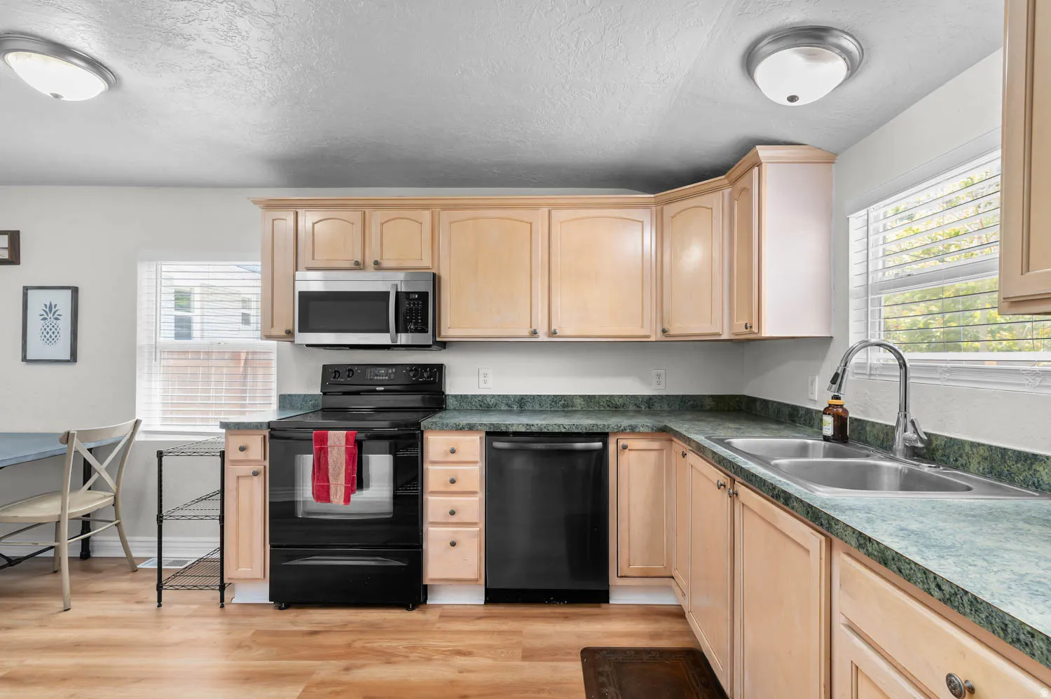Kitchen featuring a double basin stainless steel sink, light wood-finish cabinetry, black appliances including a range and dishwasher, a stainless steel microwave, and wood-finish flooring