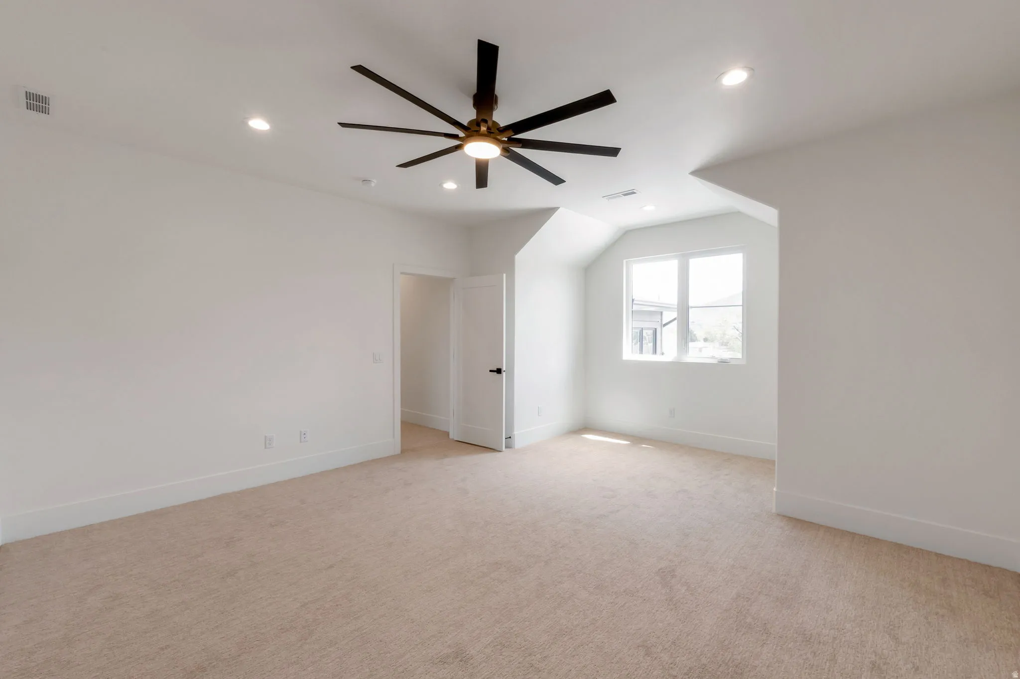 Spacious carpeted room featuring a ceiling fan with integrated lighting, recessed ceiling lighting, a window providing natural light, white baseboards, and a white interior door with dark hardware
