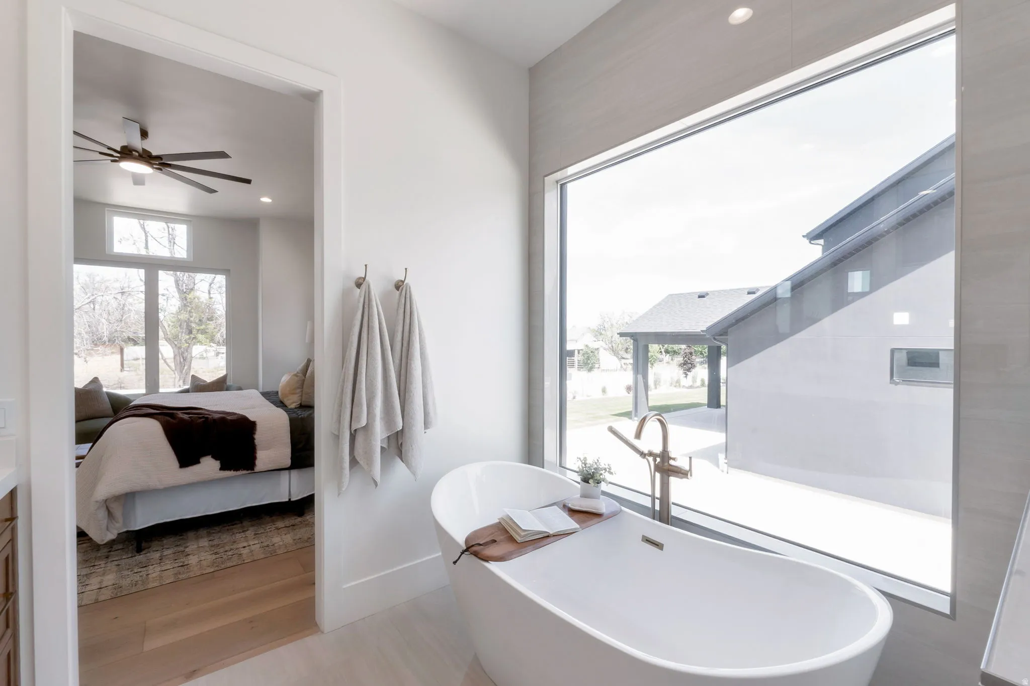 Freestanding soaking tub with gold-tone fixtures, complemented by a large picture window, light wood-finish flooring, and a neutral color palette