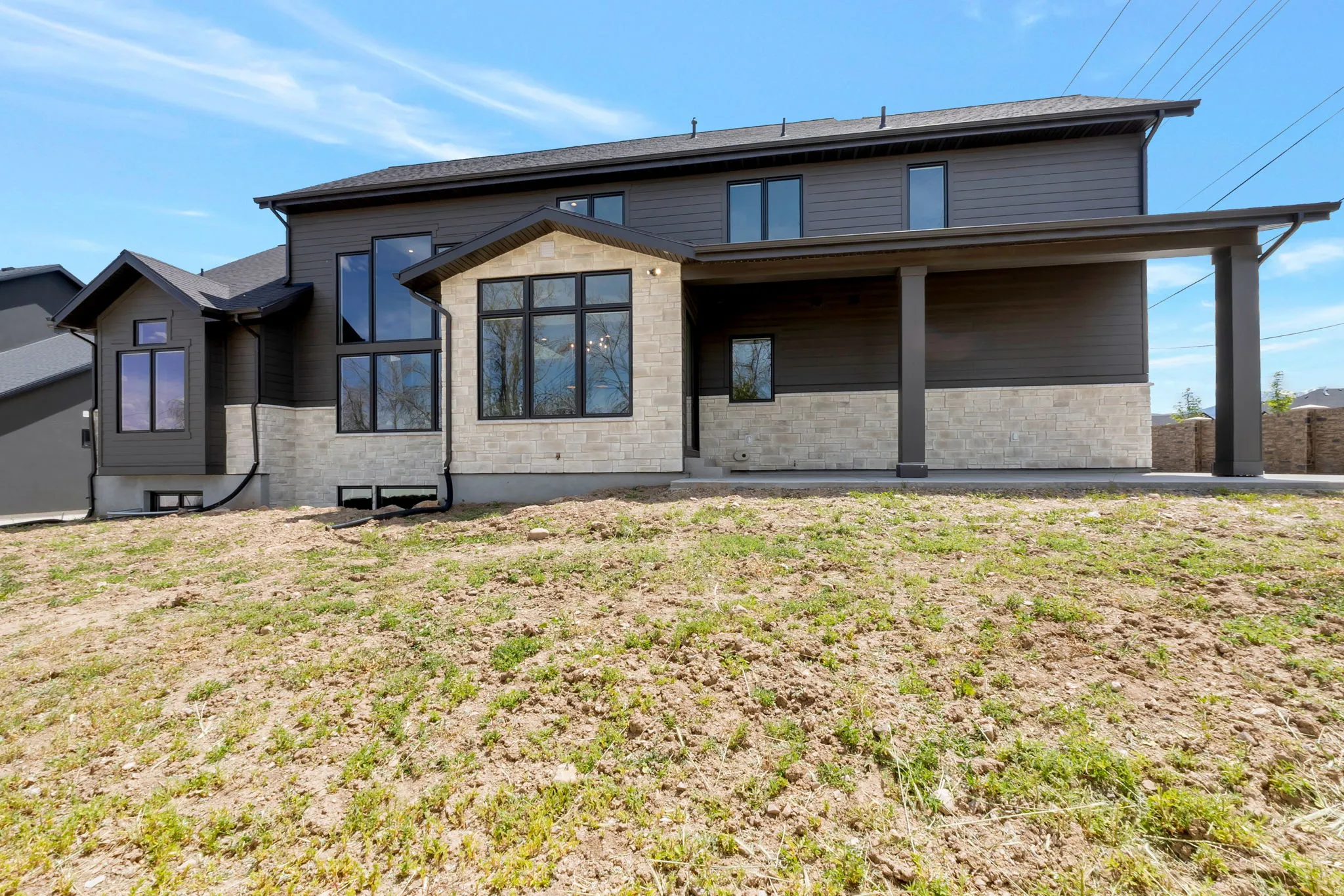 Contemporary two-story exterior featuring dark siding, stone accents, black-framed windows, a covered patio area, and a prominent multi-pane window design