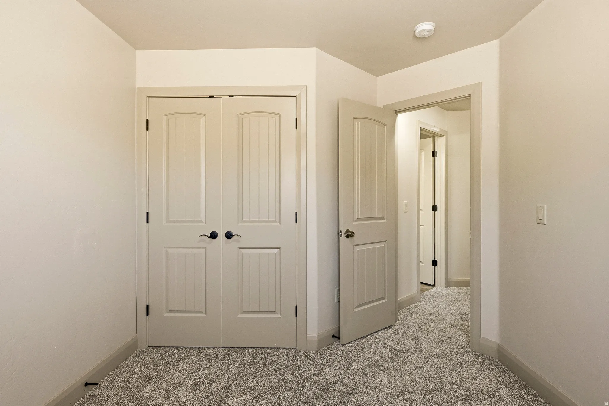 Bedroom featuring light grey carpet, white walls, white paneled bifold closet doors with black hardware, and an open doorway with a white paneled door