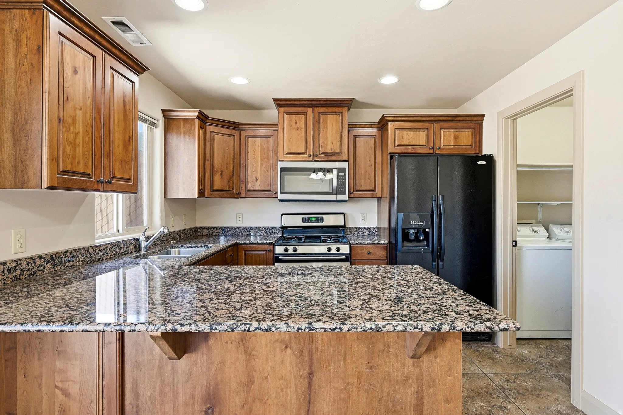 Kitchen featuring wood cabinetry, speckled countertops, stainless steel microwave, gas range, and a black refrigerator