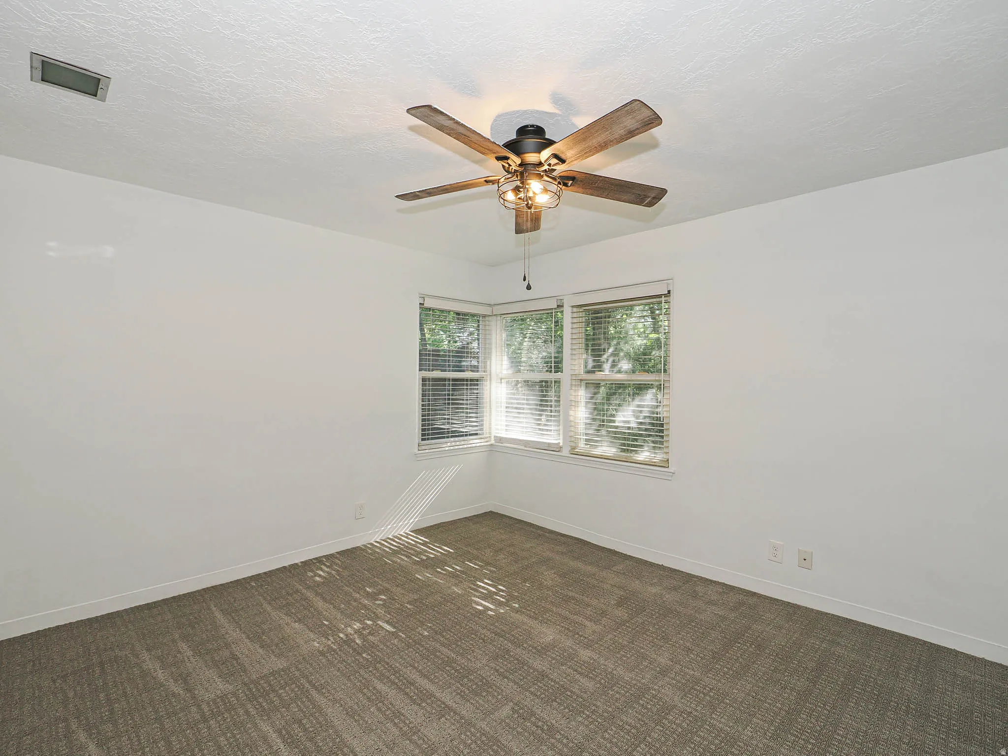 Neutral-toned room featuring a corner window arrangement with blinds, textured wall-to-wall carpeting, and a ceiling fan with integrated lighting