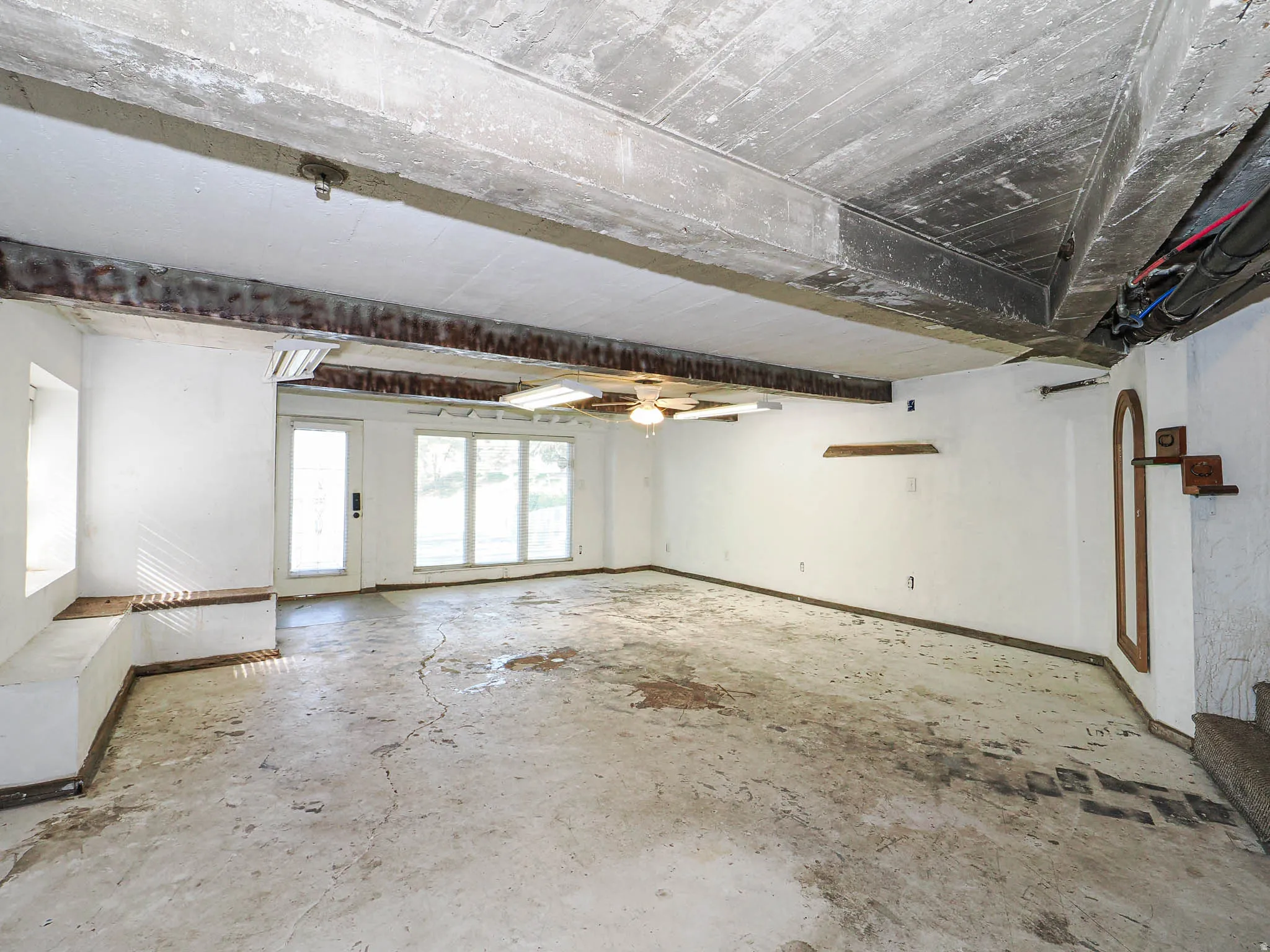 Spacious room with concrete flooring, exposed ceiling beams, a ceiling fan, and large windows with an exterior door