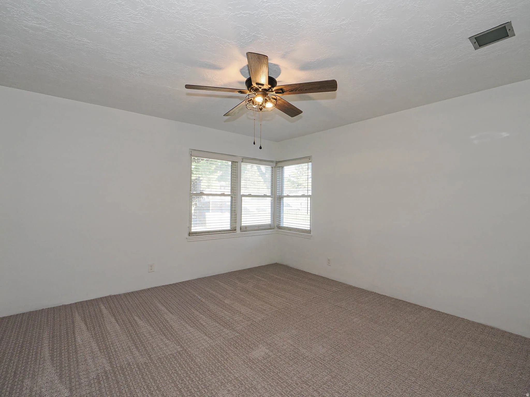 Corner window configuration with blinds, textured ceiling, brown patterned carpeting, and a ceiling fan with light fixture