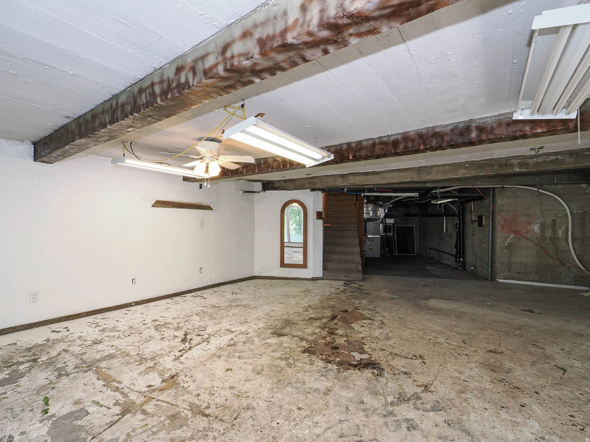 Spacious lower level offering exposed ceiling beams, concrete flooring, fluorescent overhead lighting, an arched window, and a visible staircase