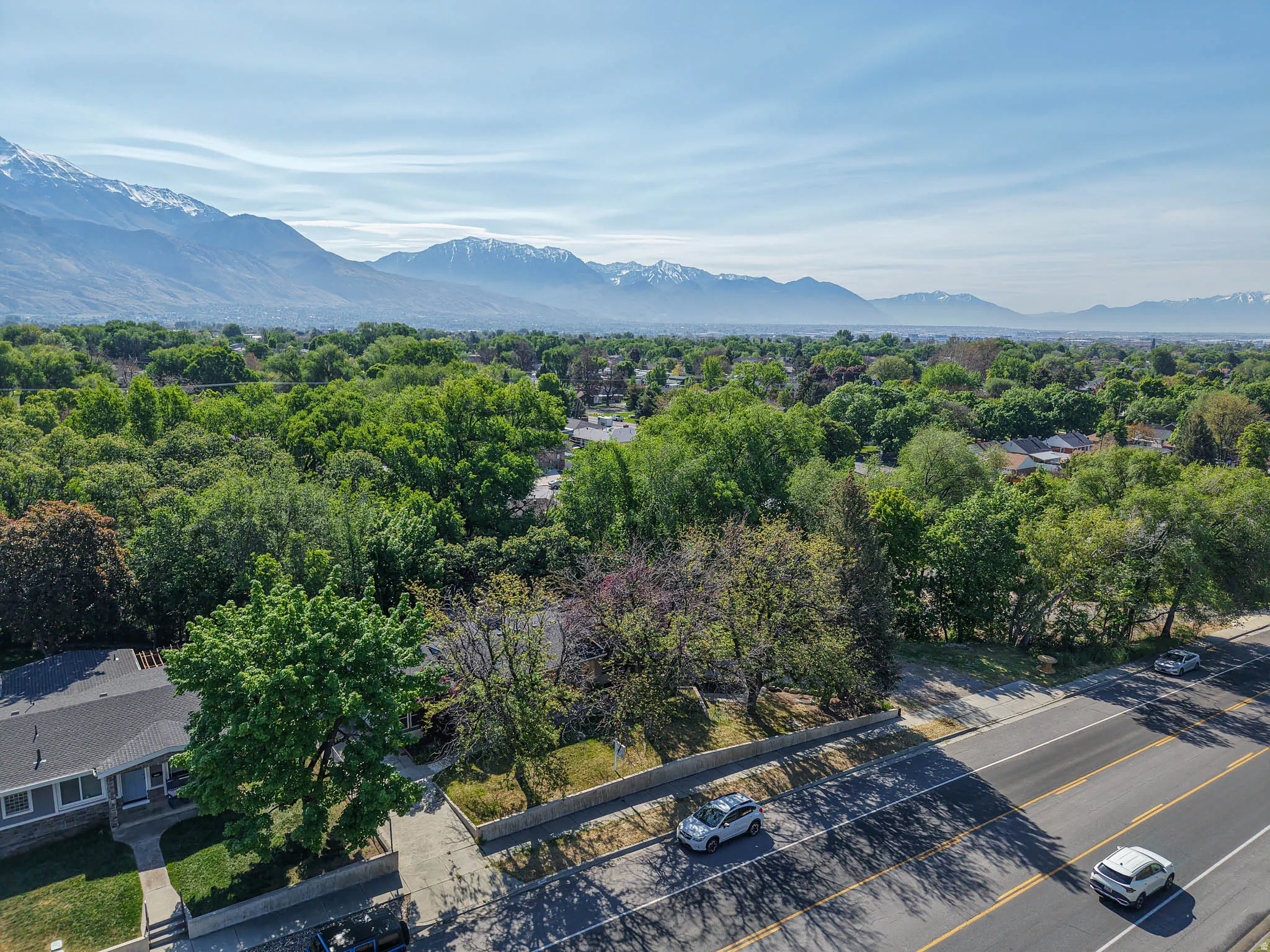 Aerial perspective showcasing a residential area with mature tree coverage, a multi-lane road, and distant snow-capped mountains