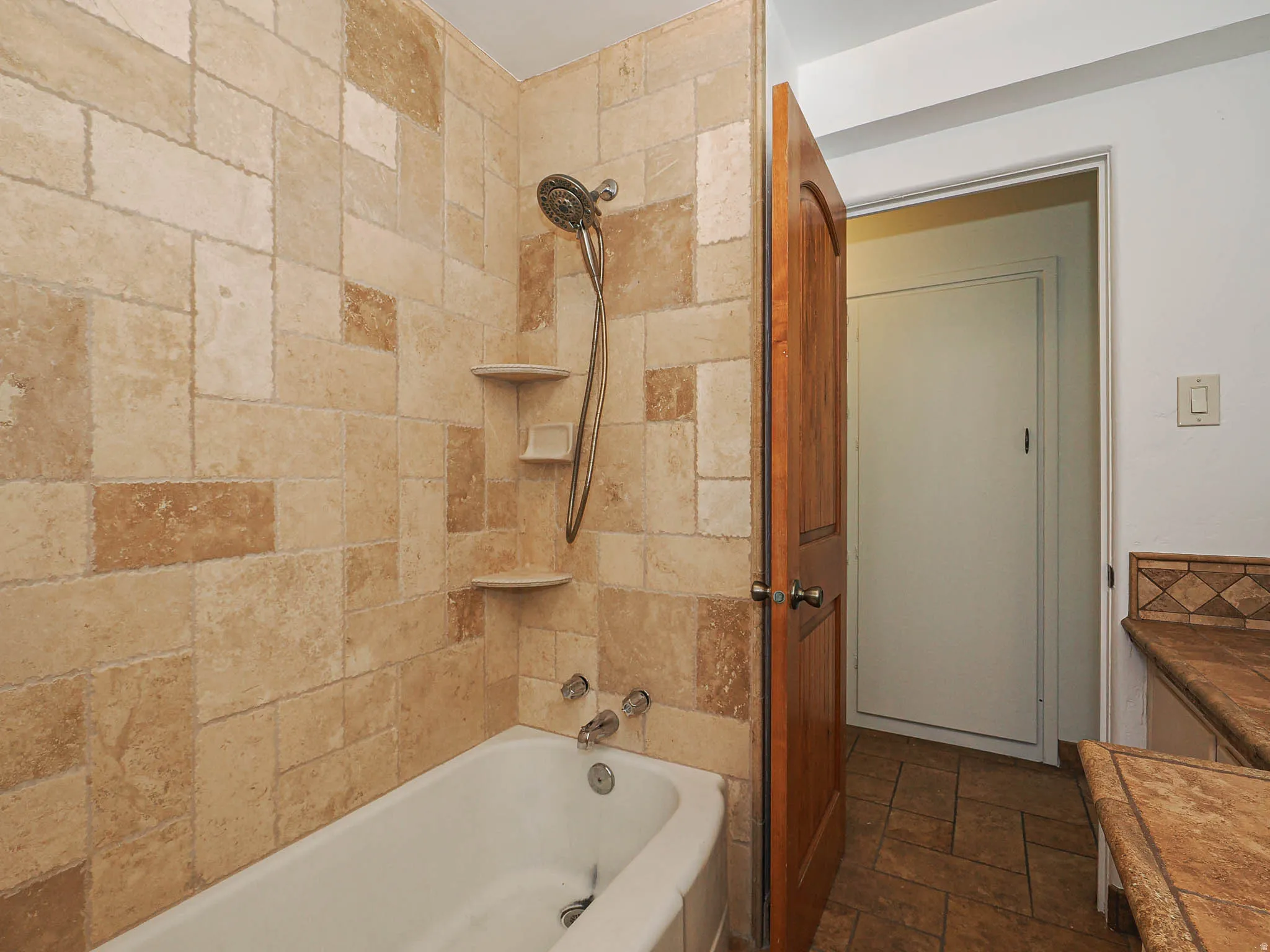 Bathtub shower combination featuring multi-toned stone tile surround, integrated corner shelves, and a handheld showerhead