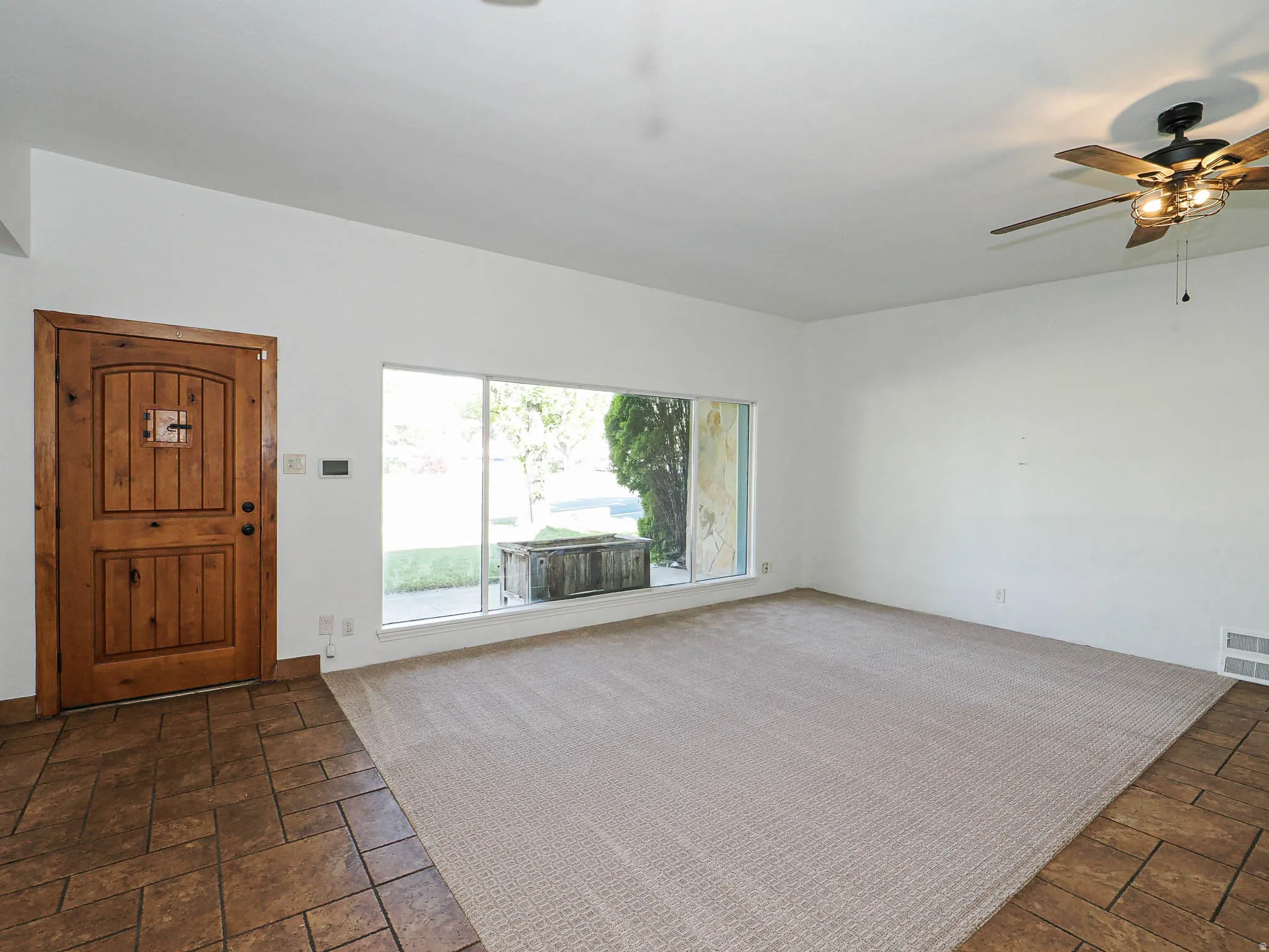Entry area featuring a solid wood door with a small glass insert, large front window, mixed flooring with carpet and tile, and a ceiling fan with integrated lighting