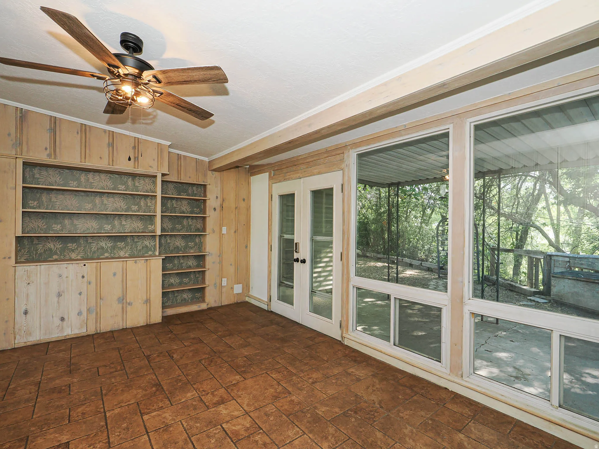 Room featuring expansive windows with exterior access, integrated shelving, wood-paneled walls, a ceiling fan, and textured tile flooring