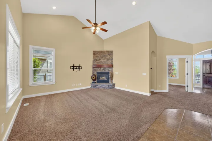 Spacious living area featuring a vaulted ceiling with a ceiling fan, a stone-faced fireplace, large windows with blinds, and carpet flooring