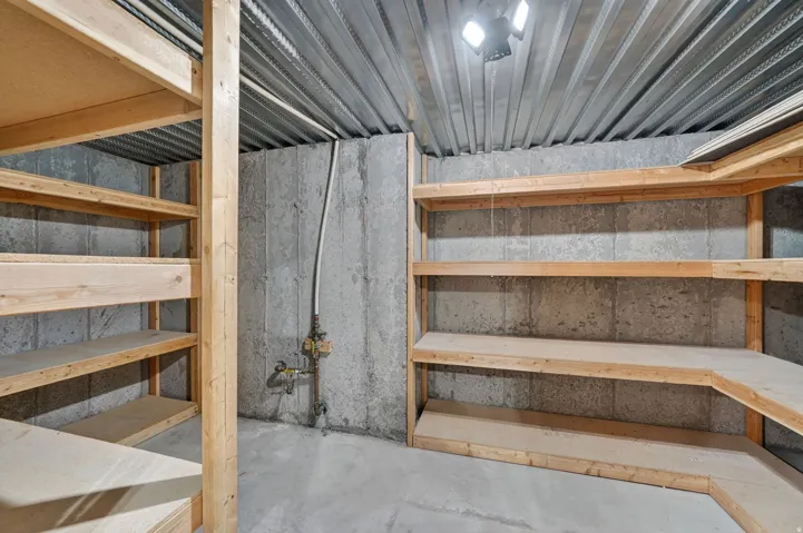 Concrete walled storage area featuring extensive built-in wooden shelving, a corrugated metal ceiling, and overhead lighting