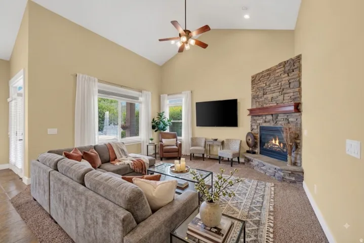 Vaulted ceilings with a ceiling fan, stone-clad fireplace with wood mantel, multiple windows, and recessed lighting