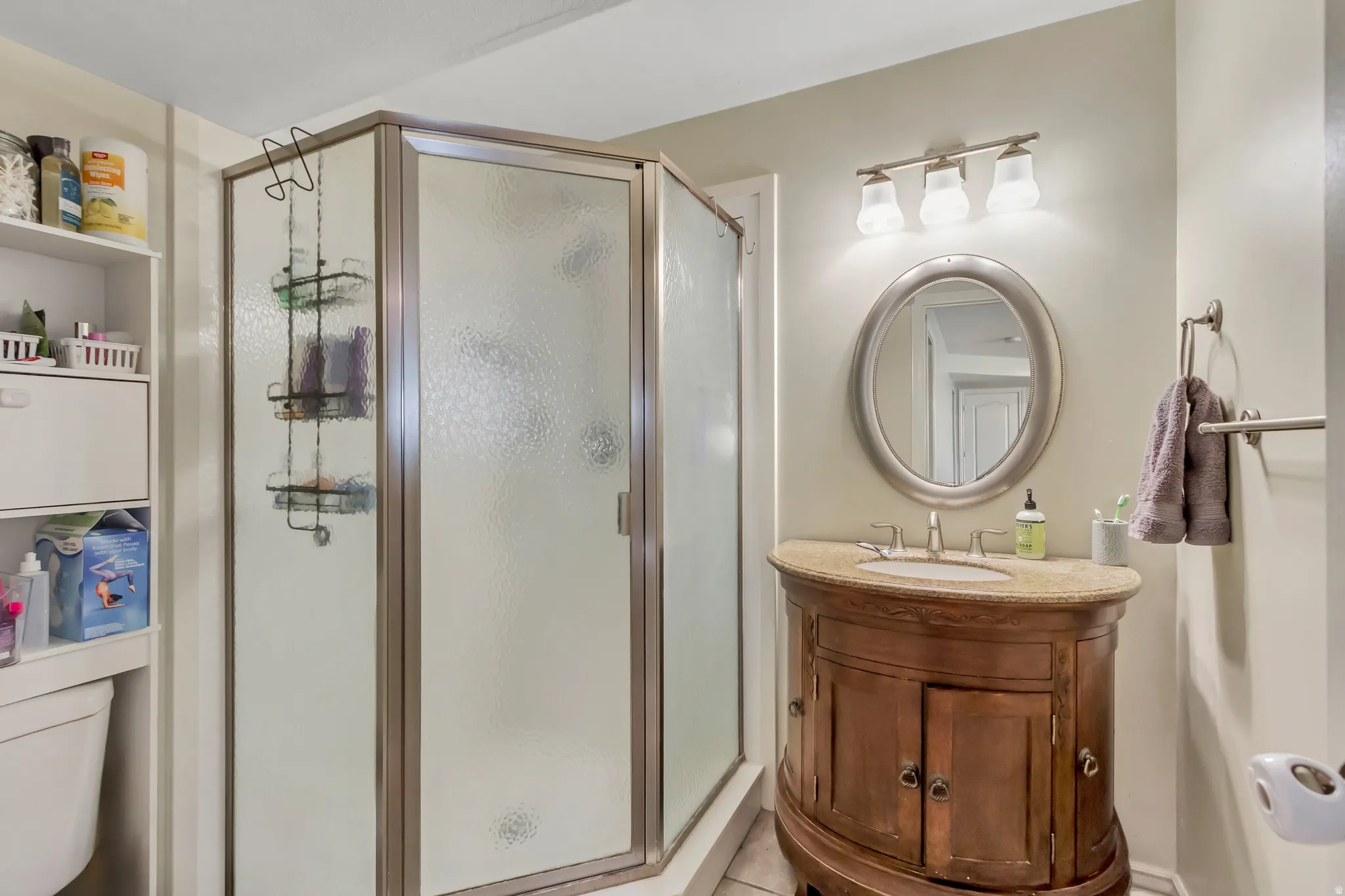 Corner shower enclosure with textured glass panels, a curved wood vanity with a stone countertop, an oval mirror, and a three-light vanity fixture