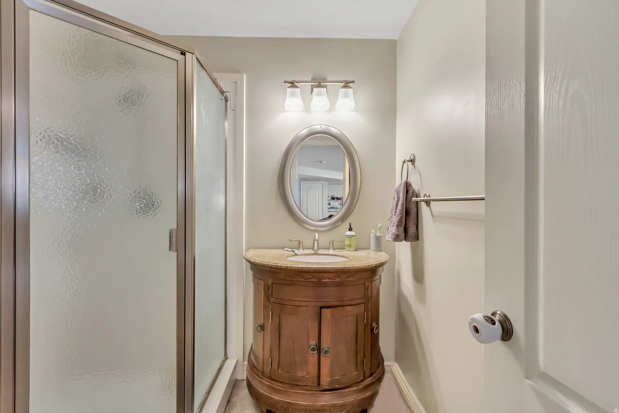 Bathroom featuring a glass-enclosed shower with textured privacy glass, a wood-finish vanity with a stone countertop, an oval mirror, and a three-light fixture