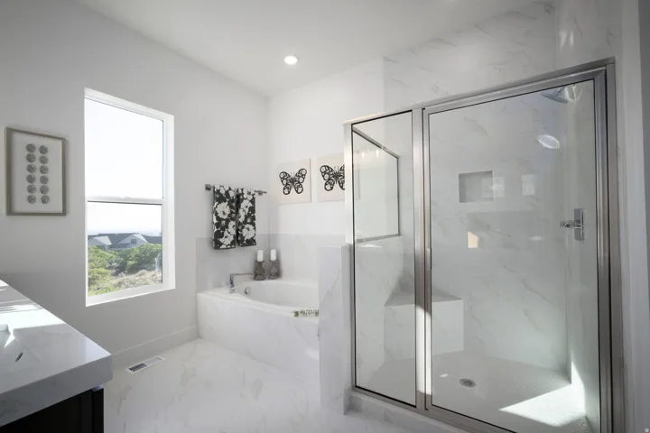 Spacious bathroom featuring a glass-enclosed shower with a built-in bench and recessed shelf, a large soaking tub, a window overlooking distant homes and greenery, white marble-look tile flooring, and a vanity with an integrated sink and light-toned count