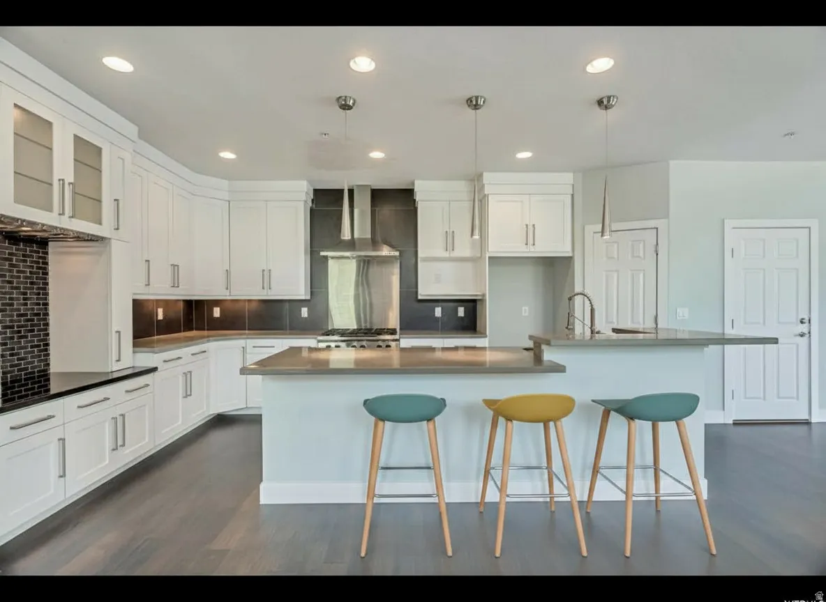 Modern kitchen featuring extensive white cabinetry, a large kitchen island with counter seating, a stainless steel range hood, dark tile backsplash, and wood-finish flooring