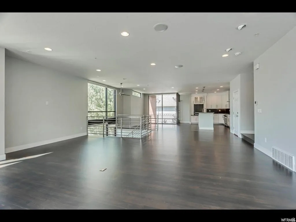 Spacious open-concept living area featuring dark wood-finish flooring, ample natural light from extensive windows, a contemporary metal railing system, recessed lighting, and a kitchen with white cabinetry